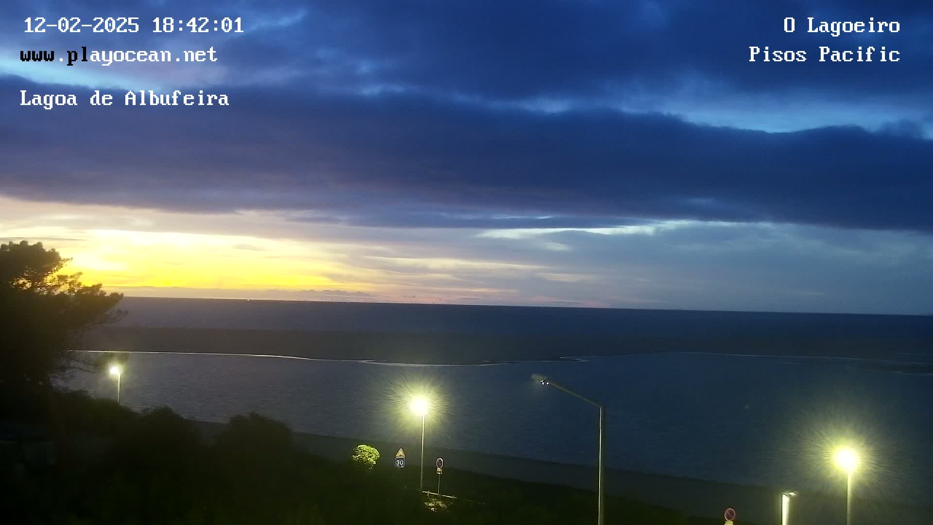 A tranquil sunset over a calm body of water, illuminated by several streetlights on a nearby shore, under a partly cloudy sky.