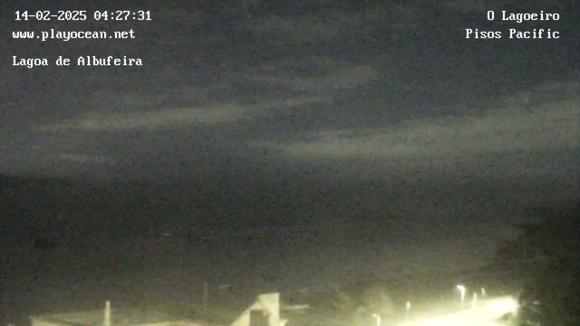 A nighttime view of a mostly cloudy sky over a dark body of water, with dimly lit buildings or structures visible in the bottom right corner.
