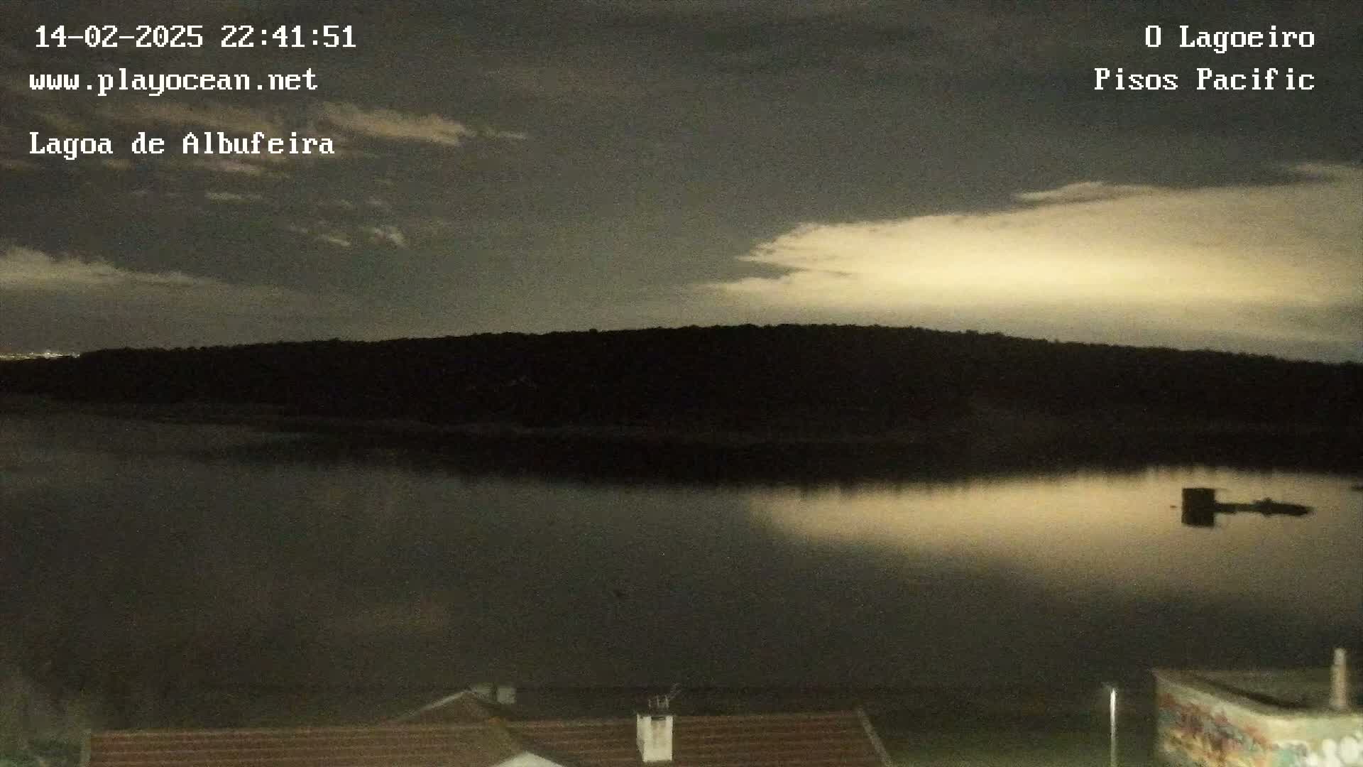 A calm body of water reflects a pale, partly cloudy night sky under which dark silhouettes of land are visible.