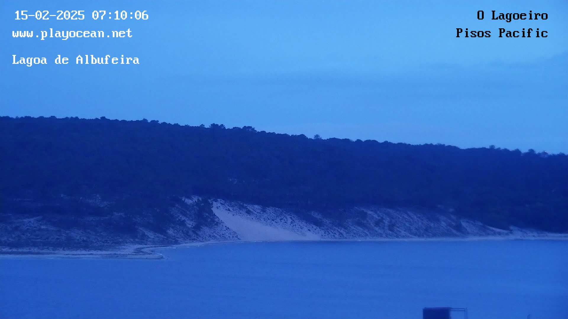 A dark blue body of water stretches out before a landmass where a sandy dune meets a dark, tree-covered hill under a clear, twilight sky.