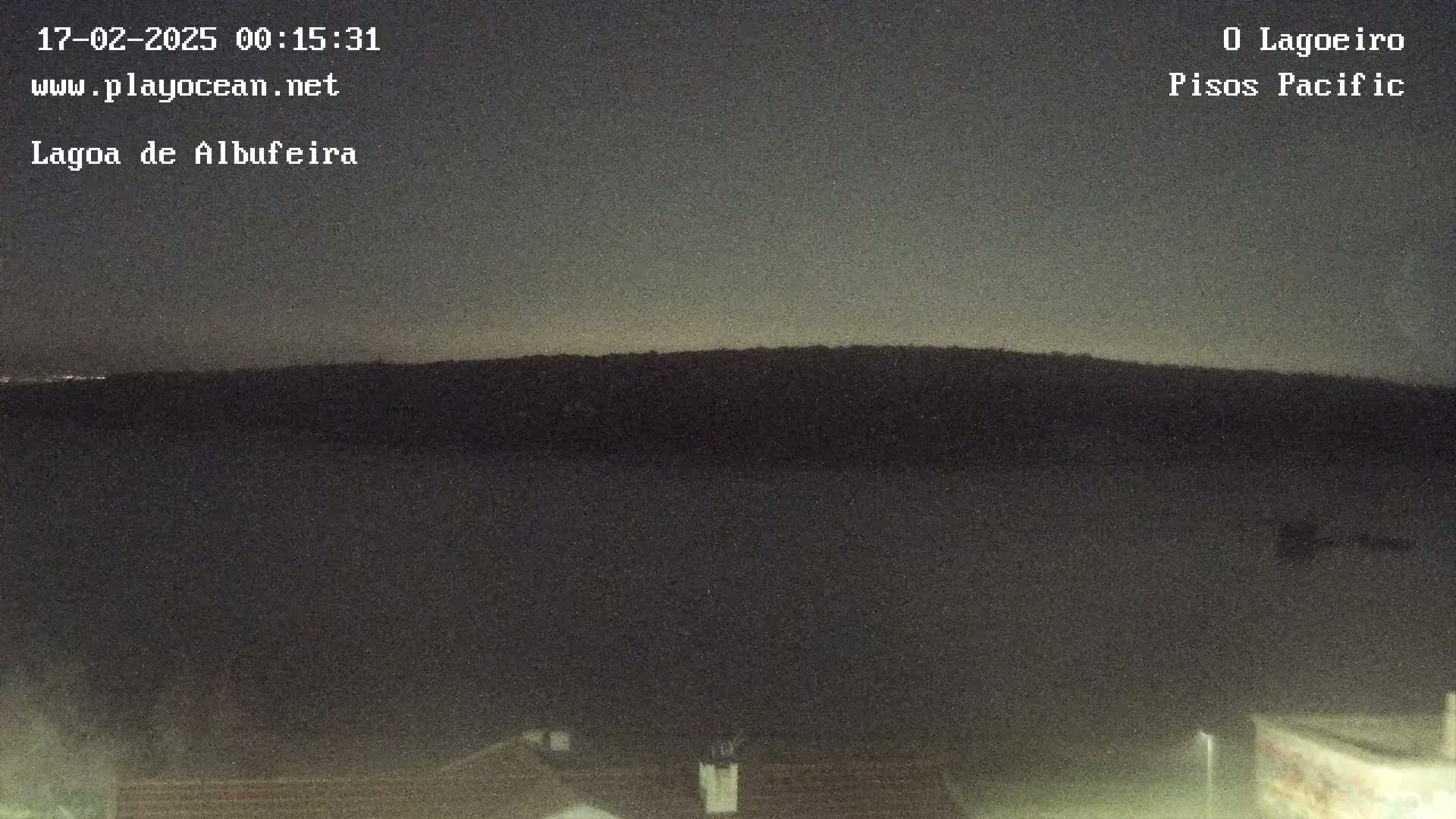 A dark, nighttime view shows a calm body of water in front of a silhouetted hill under a mostly clear sky.