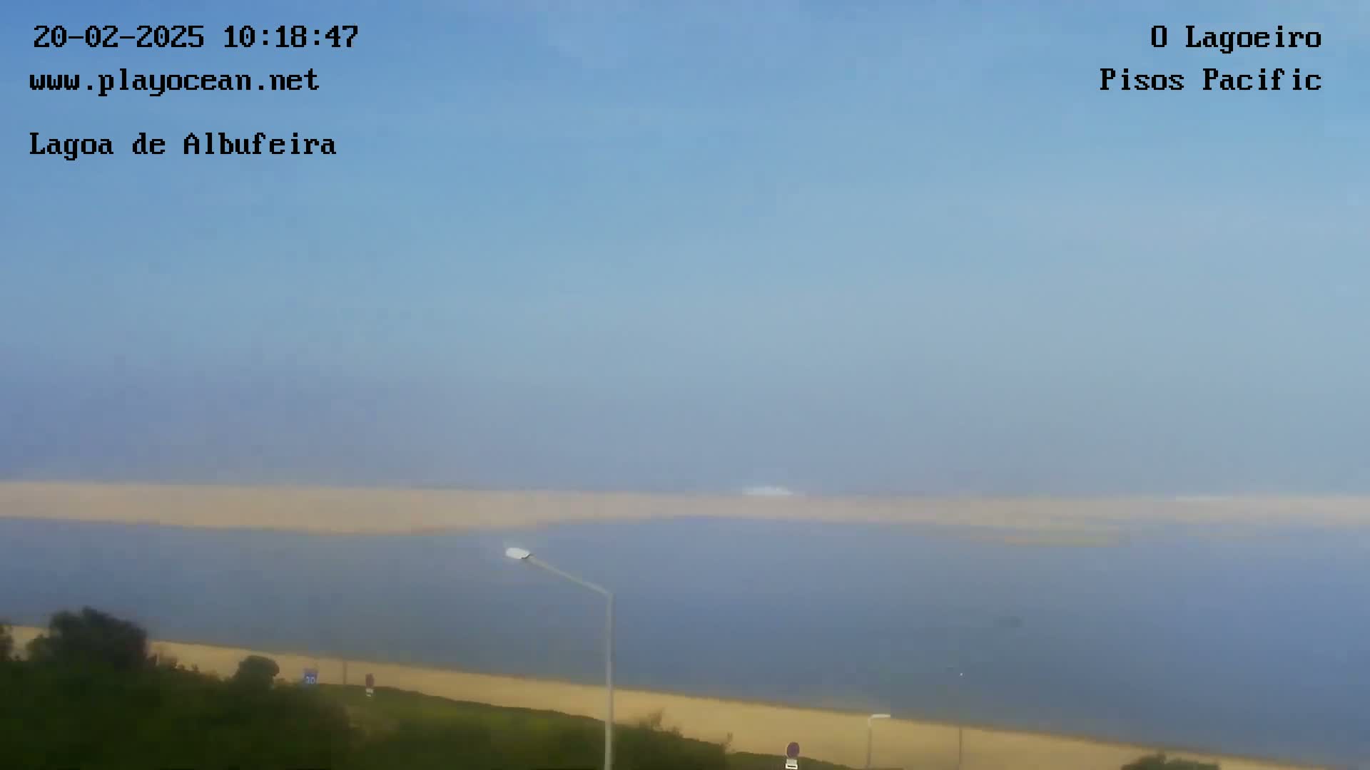 A hazy view shows a body of water with a sandy shore and some vegetation in the foreground under a mostly clear sky.