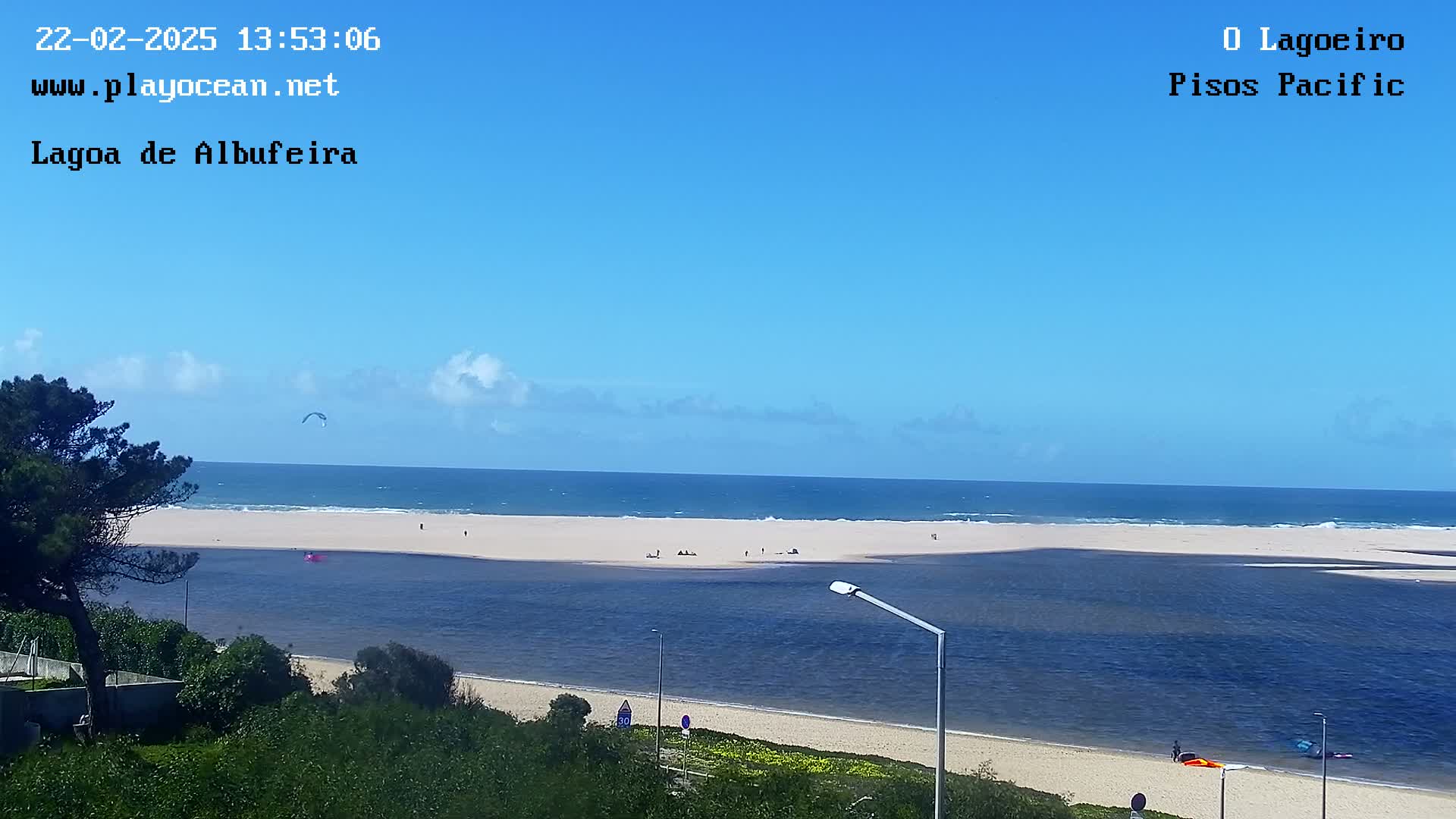 A beach scene under a clear blue sky shows a sandy beach meeting a calm body of water, with a kitesurfer in the distance.