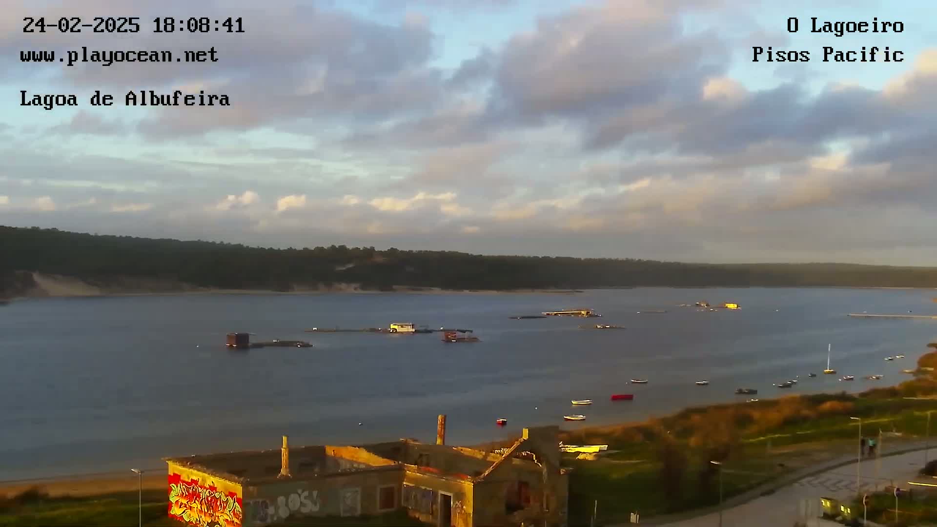 A partly cloudy evening sky overlooks a calm body of water with several small boats and structures, and a graffiti-covered ruined building in the foreground.