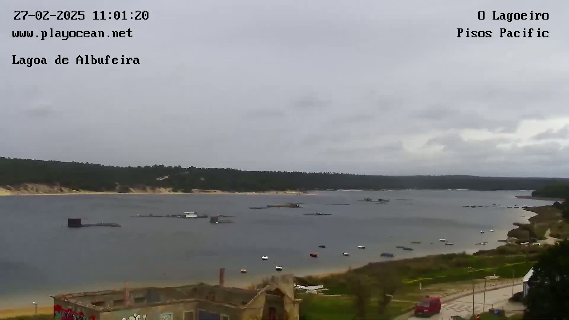 A calm, overcast day reveals a large body of water with several boats and structures, surrounded by a sandy shoreline and a green tree-lined horizon.