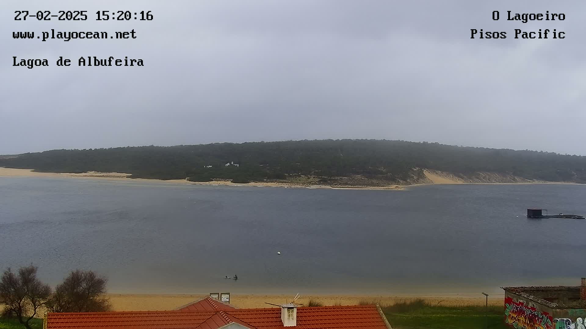 A tranquil lagoon with a sandy beach and a forested hillside under an overcast sky is visible from a vantage point above a red-roofed building.