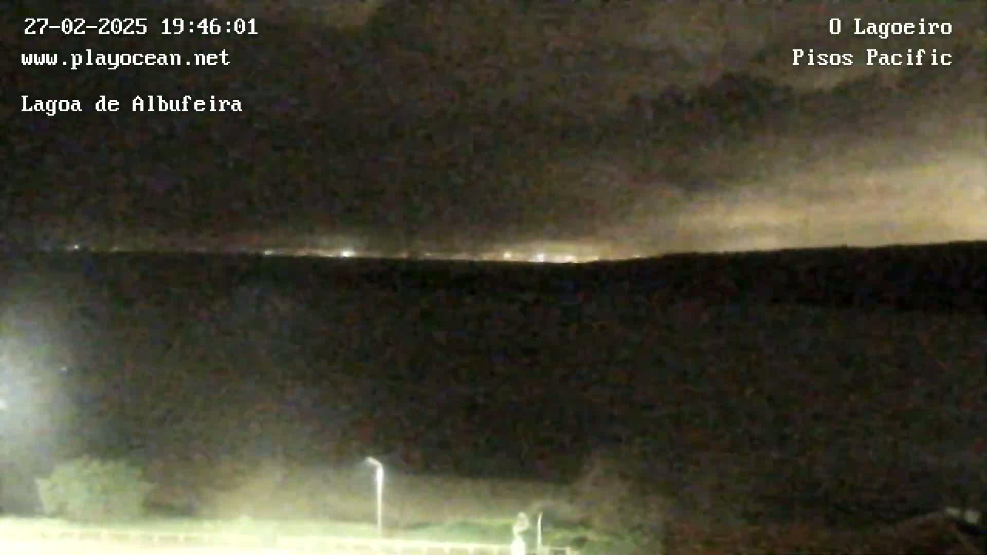 A nighttime, long shot of a dark body of water with a faint line of lights visible on the horizon under a cloudy sky.