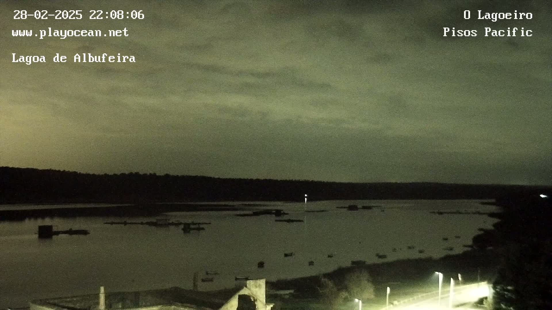 A nighttime view of a calm body of water with a few boats, surrounded by dark land under a cloudy sky.