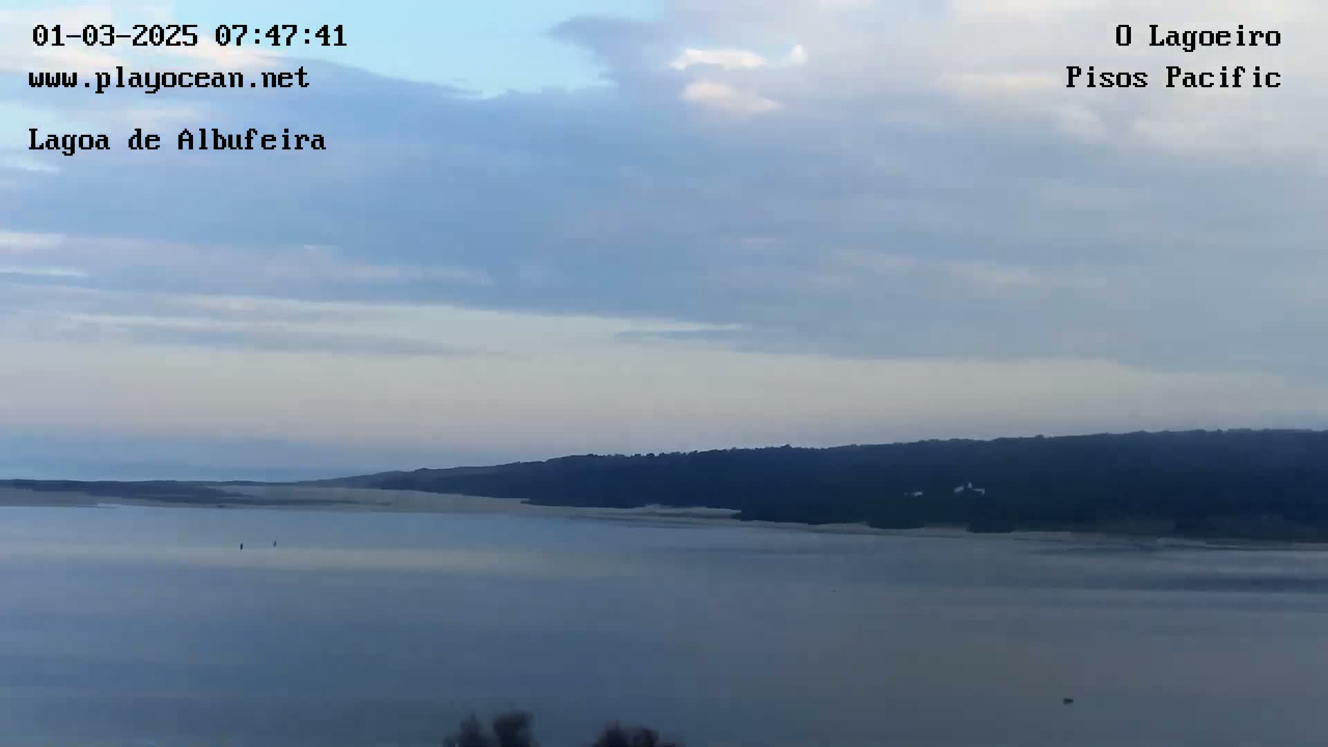 A calm body of water stretches out beneath a mostly cloudy sky, with a dark line of trees and land visible on the horizon.