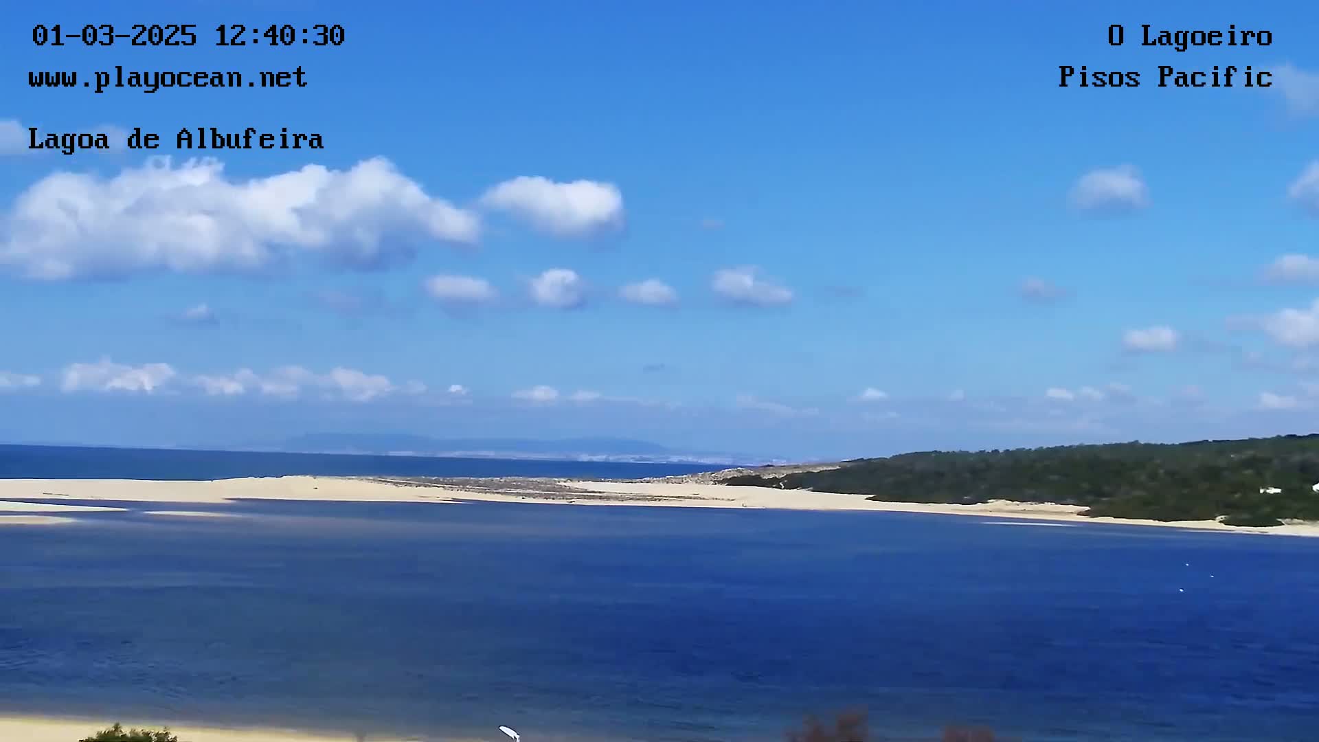 A tranquil lagoon scene under a mostly sunny sky with scattered cumulus clouds features a sandy shoreline and distant hills.
