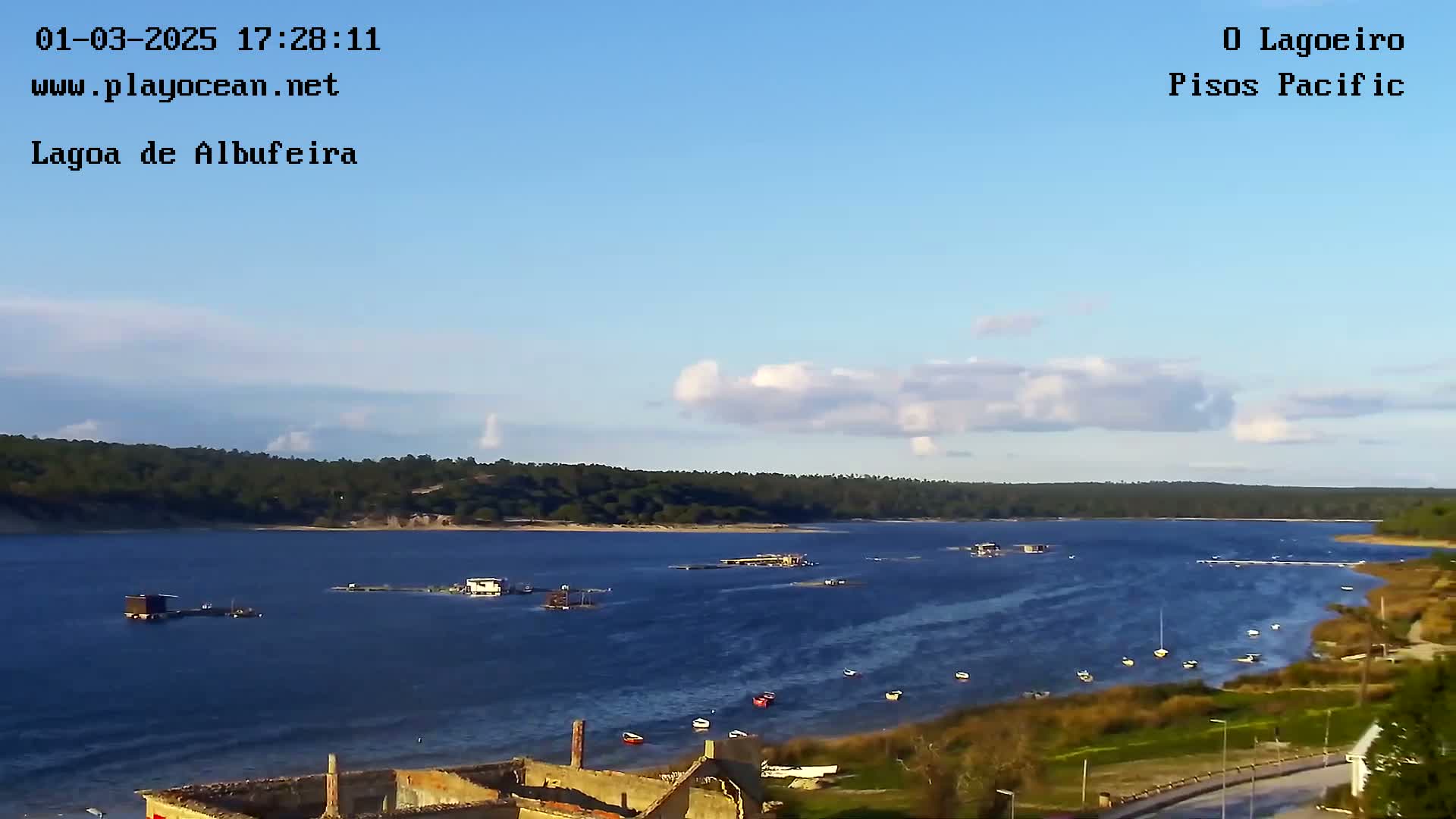 A calm body of water with scattered boats and a few structures on the shore under a partly cloudy blue sky.