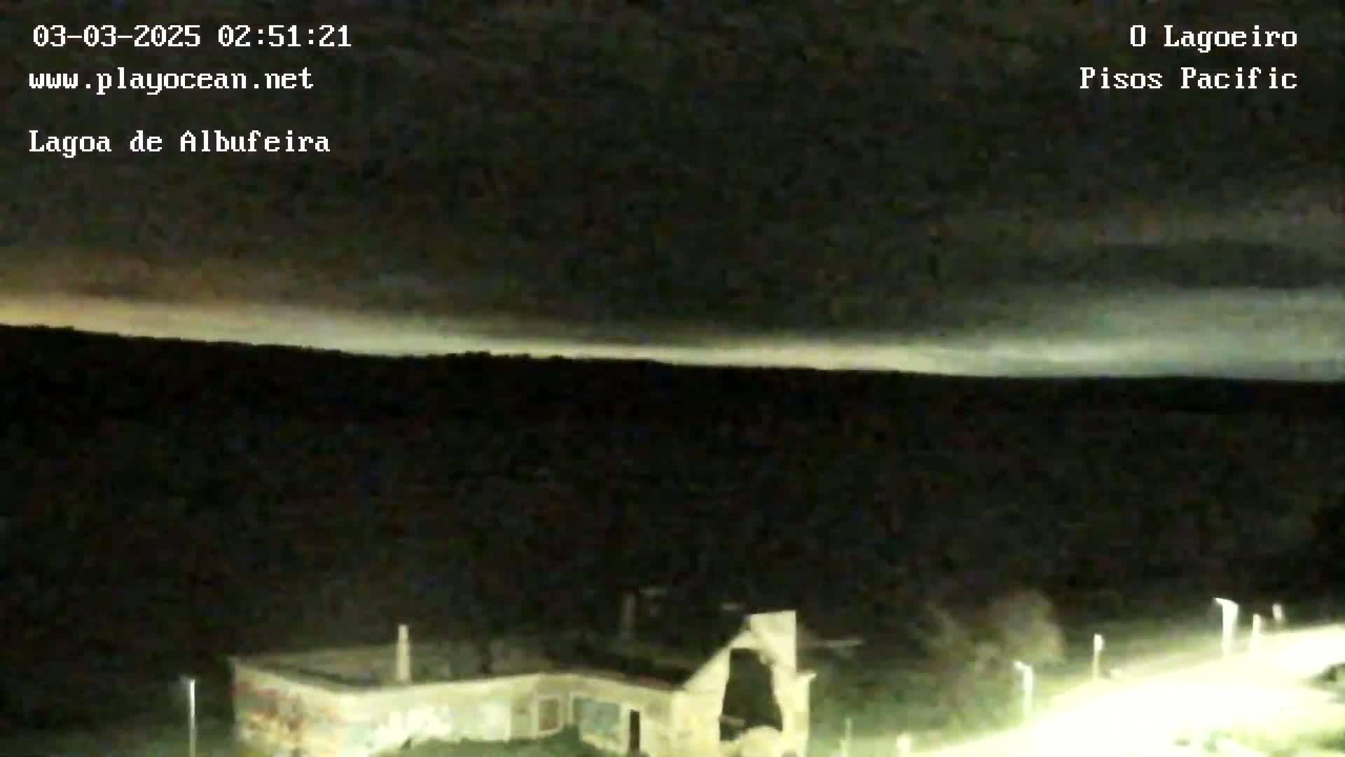 A low-light, nighttime image shows a pale, horizontal band of clouds across a dark sky above a dark landscape with some illuminated buildings below.