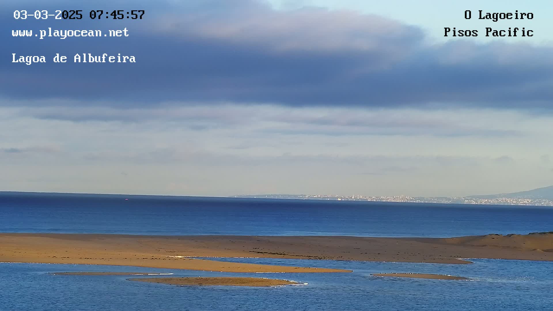 A calm ocean with a mostly cloudy sky, showing a sandy shoreline with small inlets and a distant city skyline.