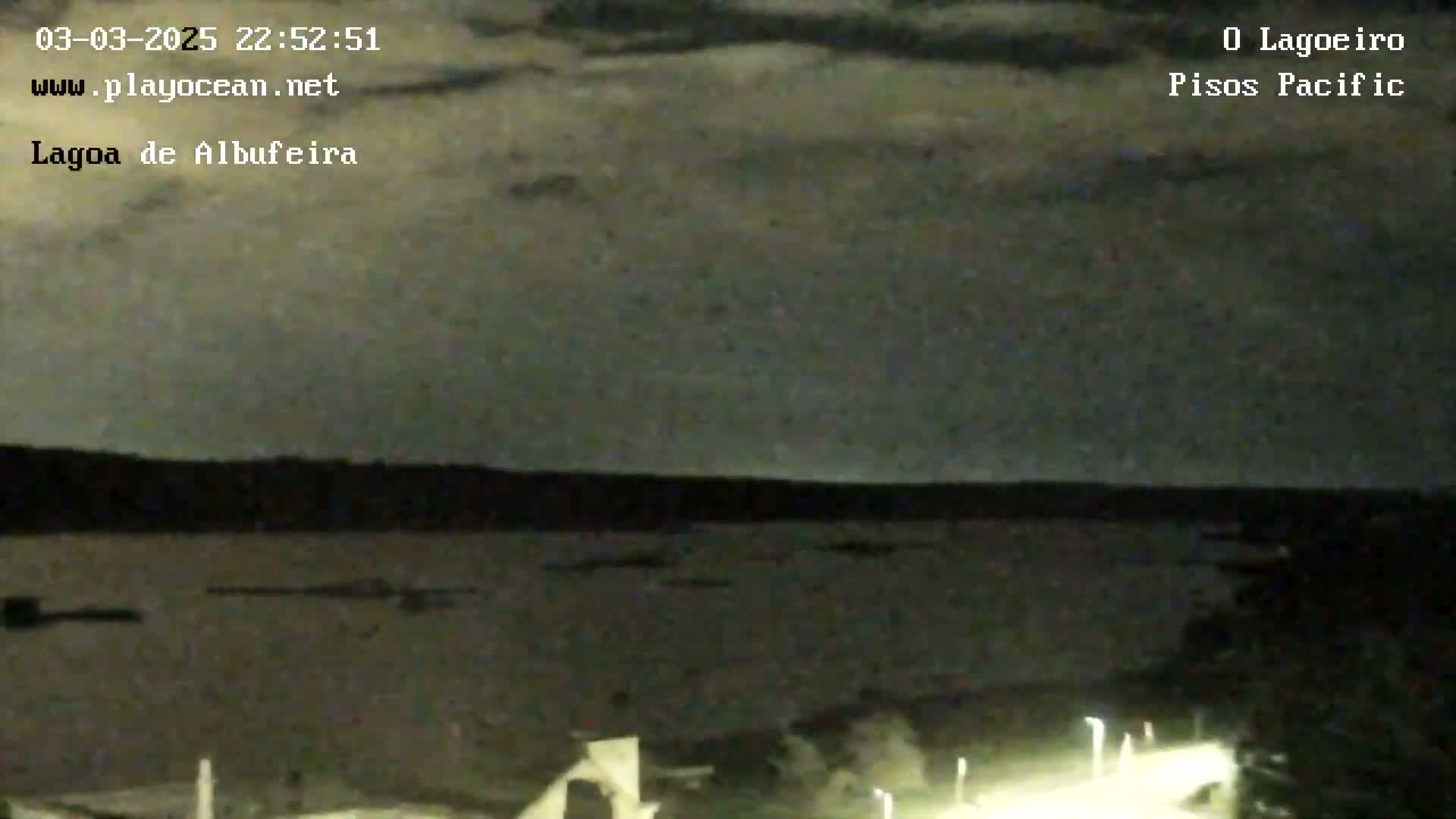 A nighttime view of a dark body of water under a cloudy sky shows a shoreline with lights in the foreground.