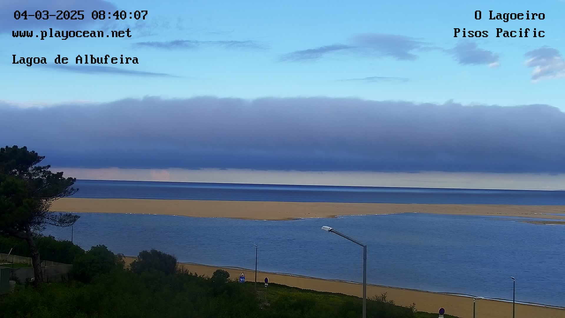 A mostly cloudy sky hangs over a calm body of water that leads to a sandy beach and ocean under a mostly cloudy sky.