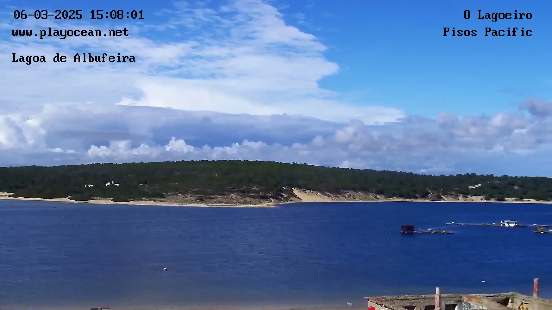 A calm body of water stretches towards a verdant, tree-covered shoreline under a partly cloudy sky.