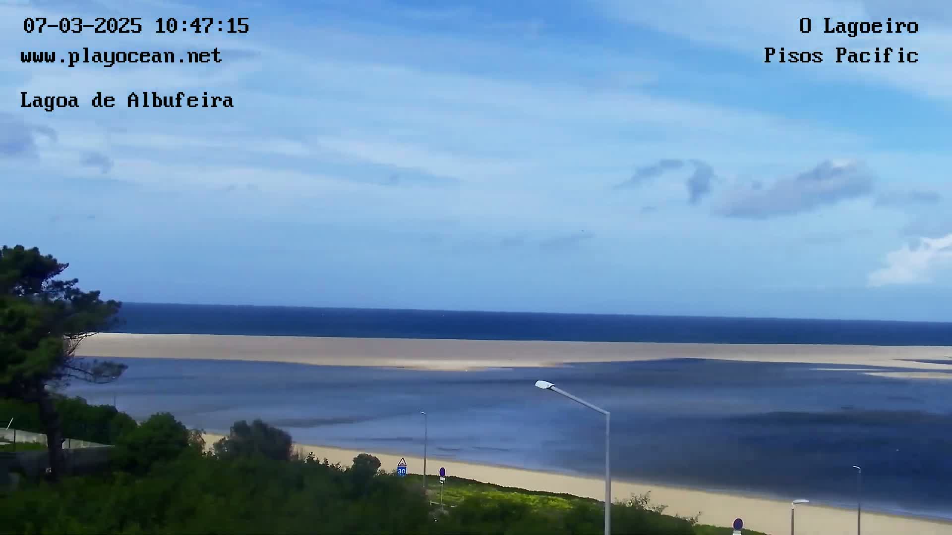 A mostly sunny day overlooks a tranquil lagoon that connects to a calm ocean, with green vegetation and a road visible in the foreground.