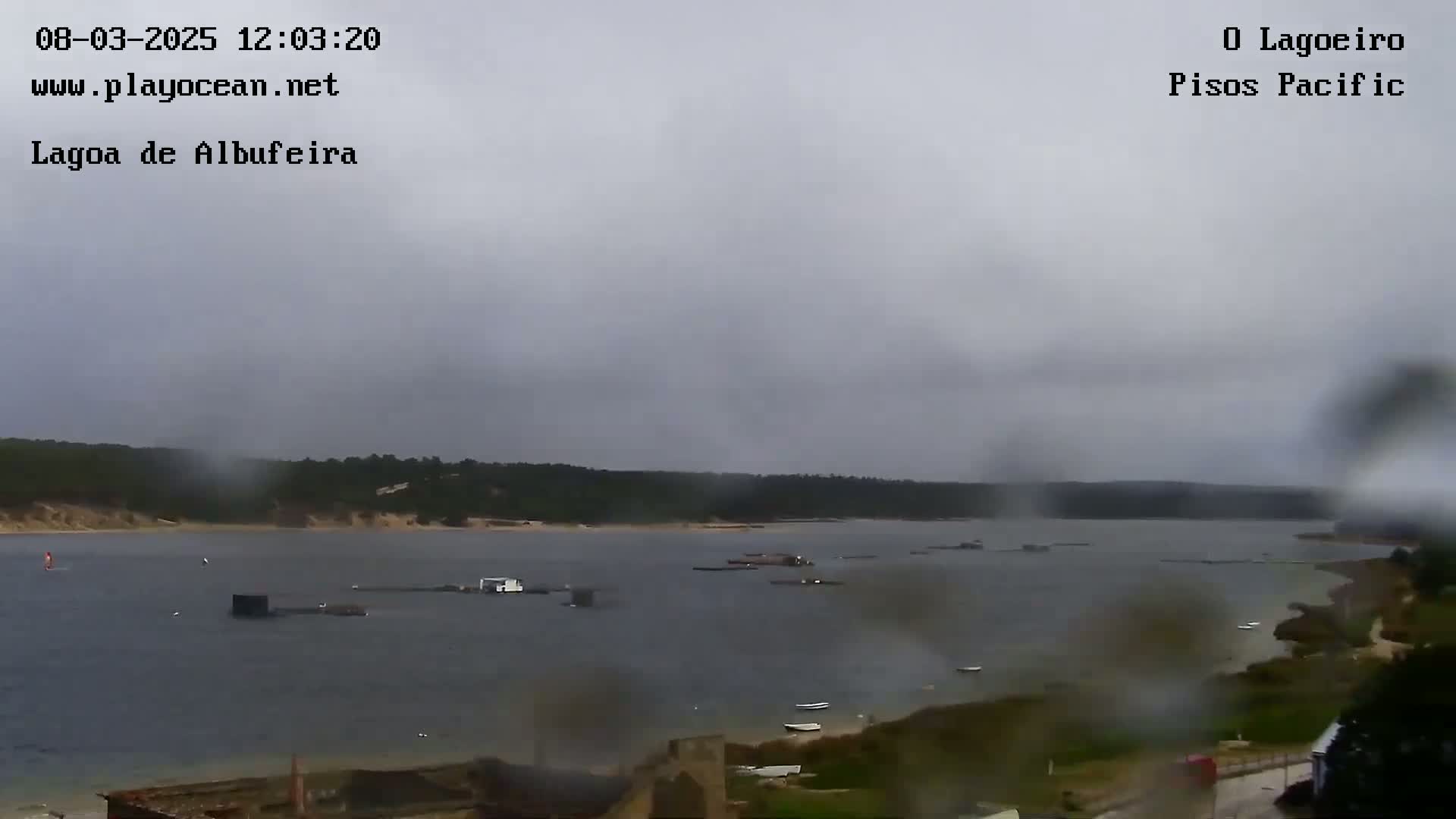 A wide shot of a body of water on an overcast day with rain, showing a shoreline with sparse vegetation and several small structures or boats on the water.