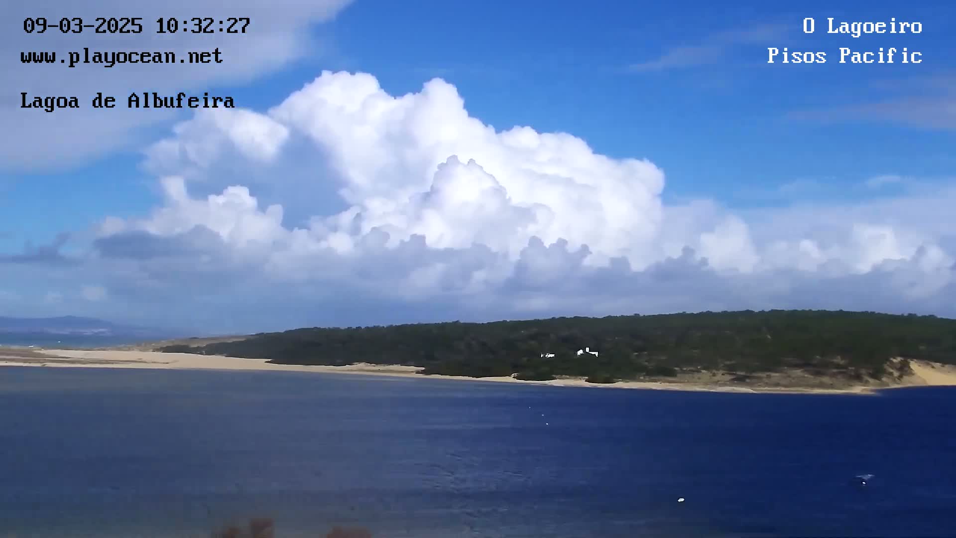 A partly cloudy day shows a large body of water with a sandy shore and green hills in the background.