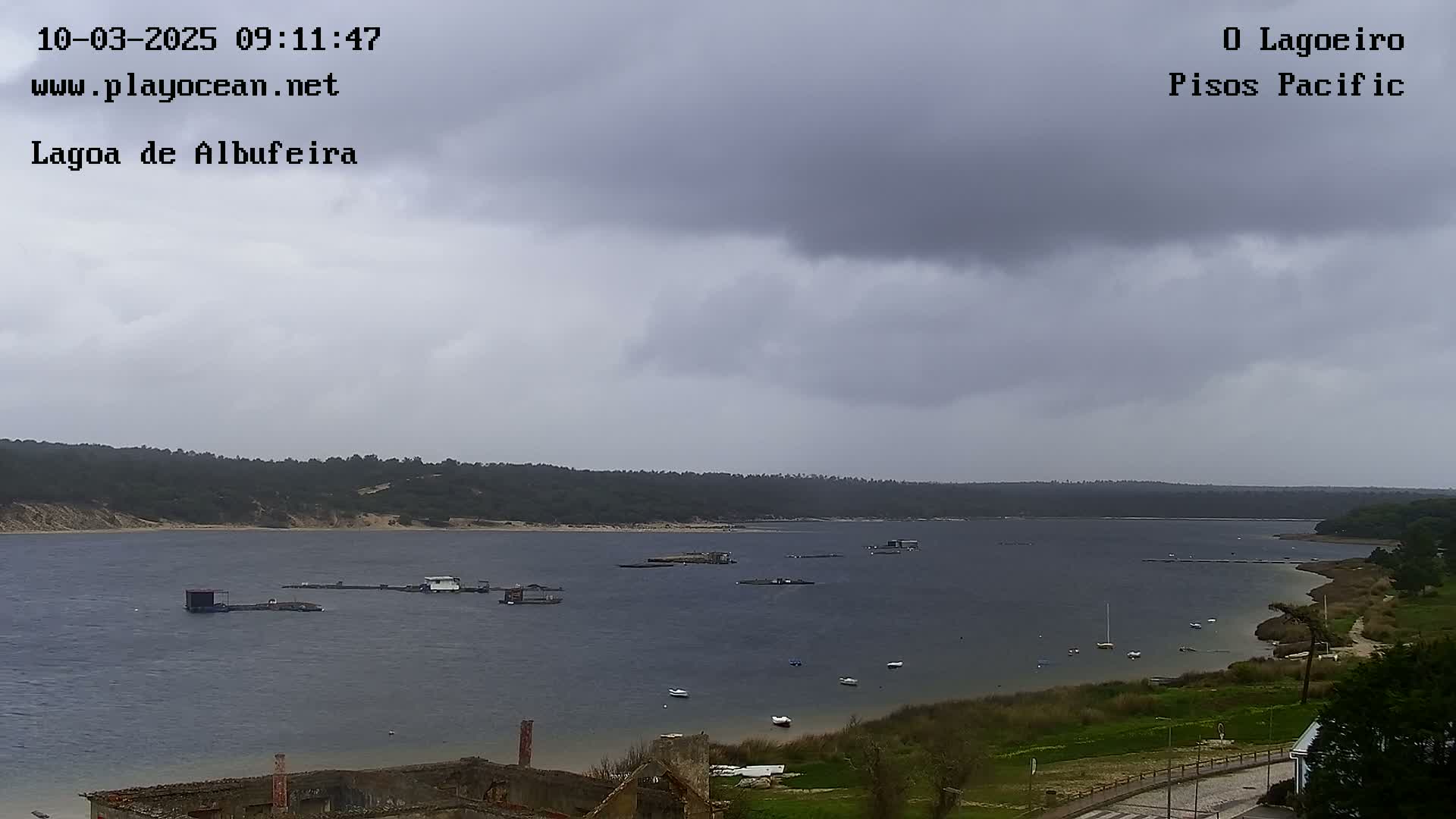 A wide shot shows a calm body of water under an overcast sky, with several small boats and structures visible on the water and a shoreline with greenery and some buildings.