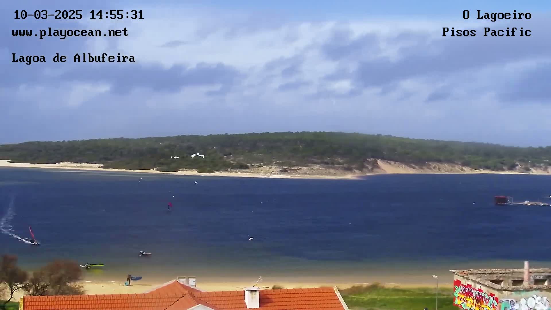 A body of water with several sailboats and windsurfers is seen from a high vantage point on a partly cloudy day, with a sandy beach and houses visible in the foreground.
