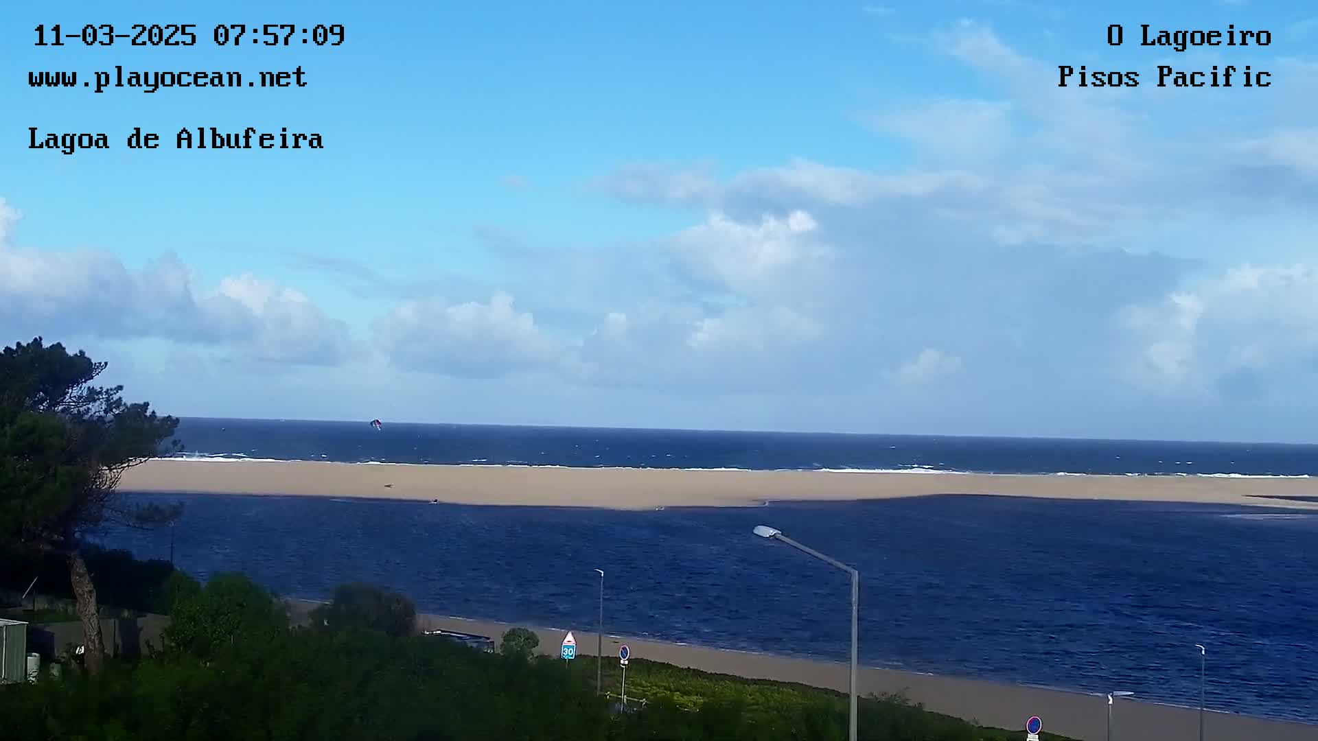 A partly cloudy day shows a wide sandy beach meeting a dark blue body of water, with a kitesurfer visible in the distance.
