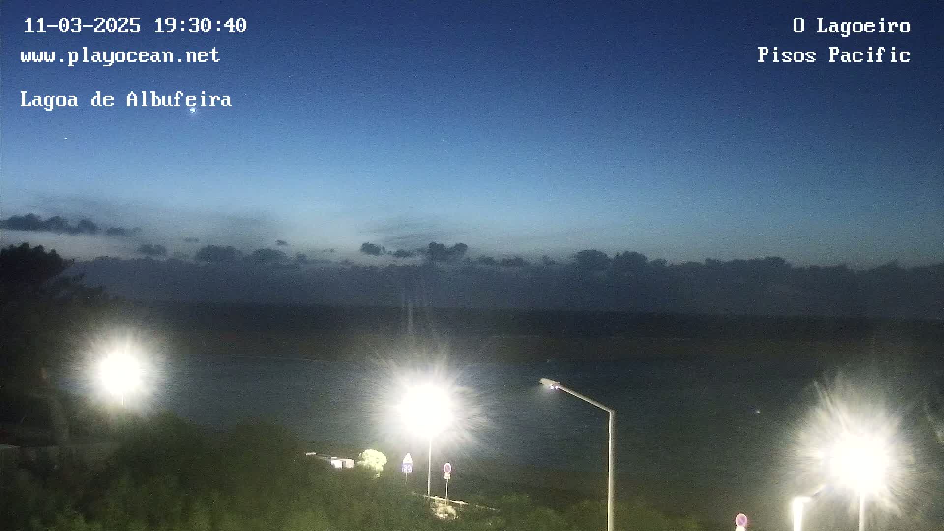 A dark-blue twilight sky with a few clouds hangs over a calm body of water, illuminated by several bright streetlights on a hillside.