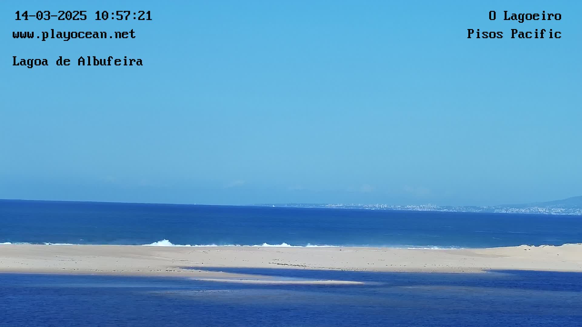 A sandy beach separates a calm lagoon from a blue ocean under a clear sky, with a distant cityscape visible on the horizon.