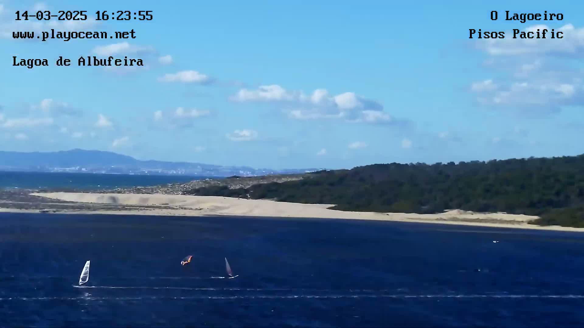 Two sailboats are on a body of water near a sandy shore backed by green hills under a partly cloudy blue sky.