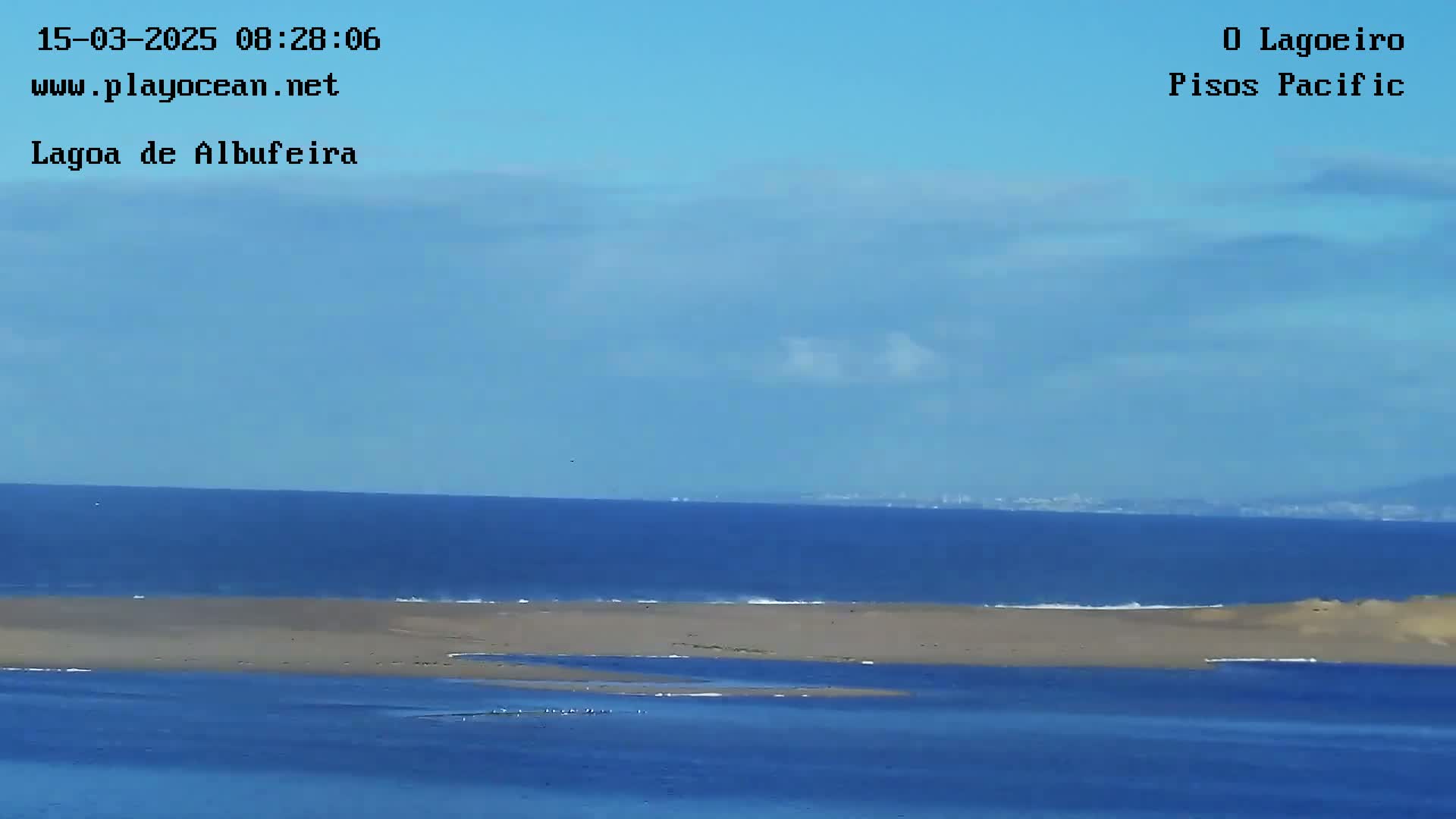 A mostly clear blue sky overlooks a calm ocean with a sandy landmass separating the ocean from a lagoon.