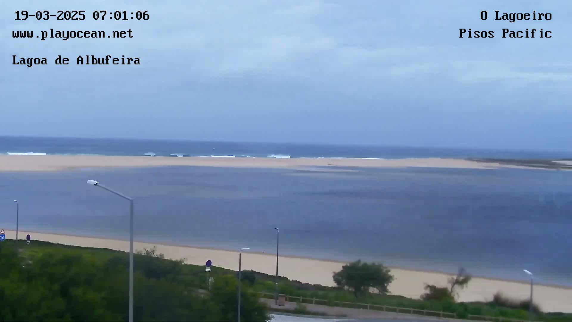 A calm ocean with small waves meets a sandy lagoon shore, viewed from an elevated position on an overcast day.