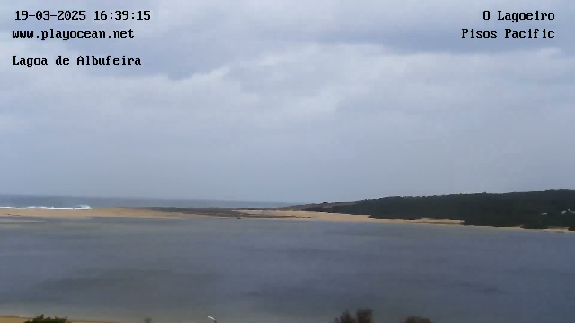 A calm lagoon with a sandy shore and dark green vegetation on the far bank is visible under a mostly cloudy sky.