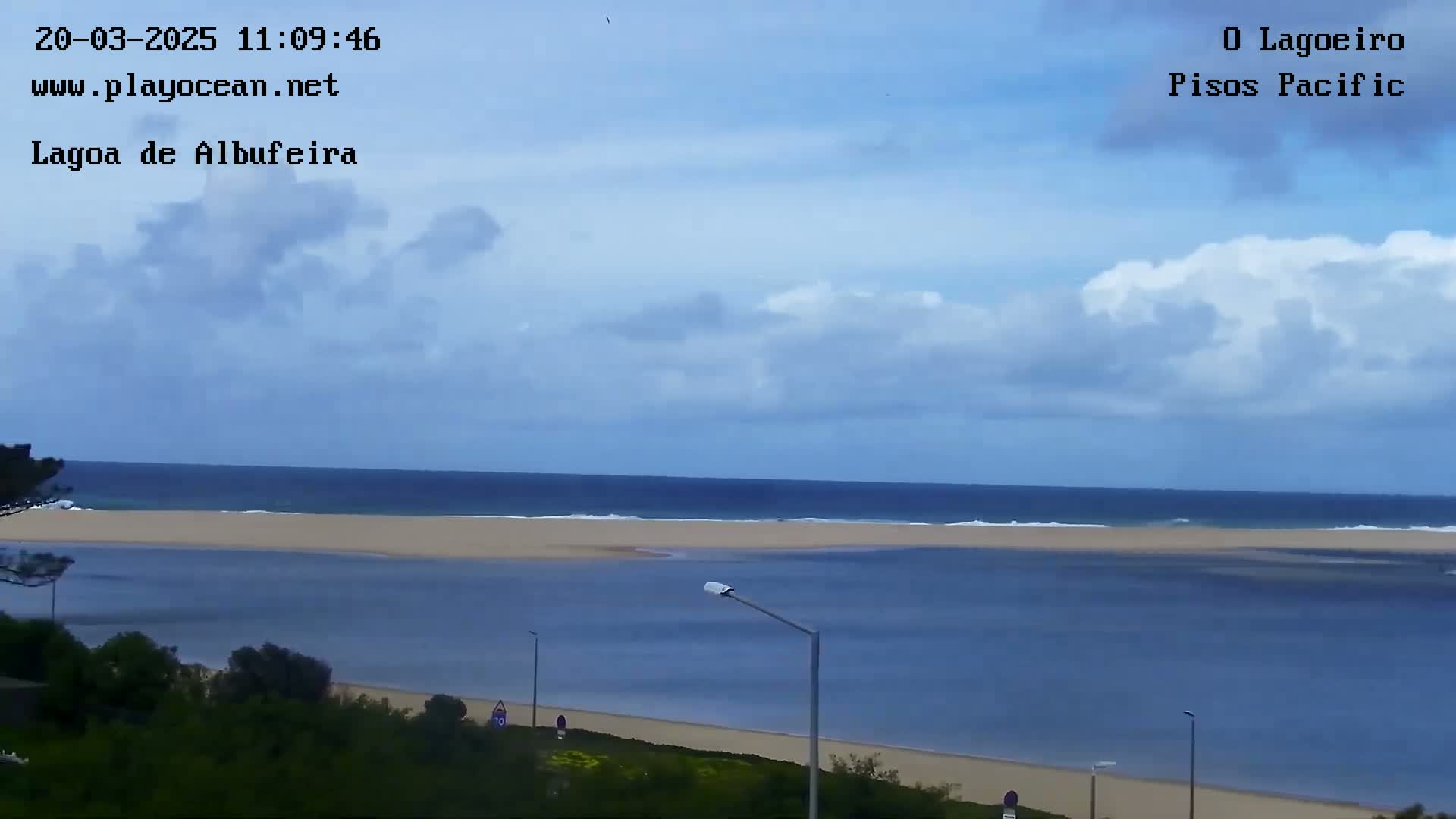 A partly cloudy day reveals a view of a calm lagoon meeting the ocean, with a sandy beach separating the two bodies of water.