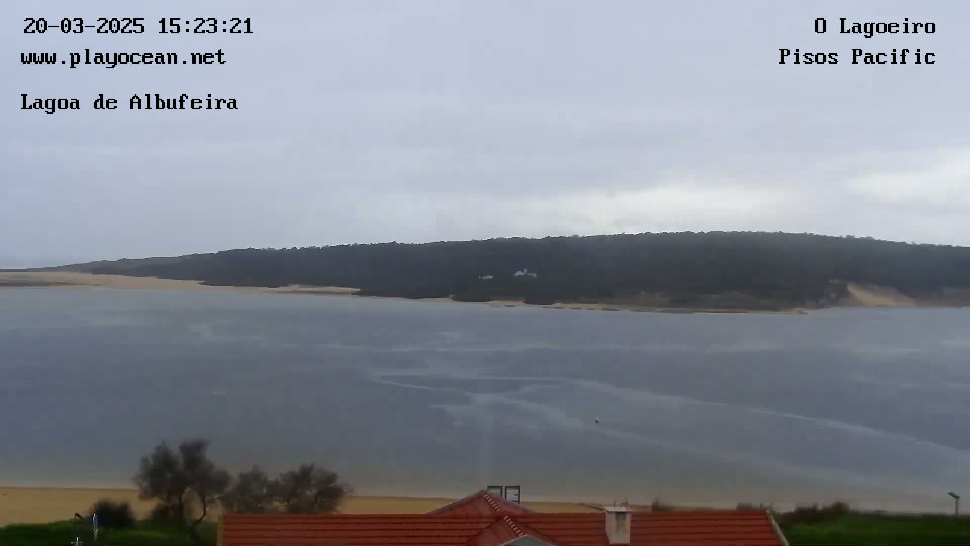 A calm, overcast day shows a large body of water extending to a sandy shore and wooded hillside in the distance, with a small portion of a red-roofed building visible in the foreground.