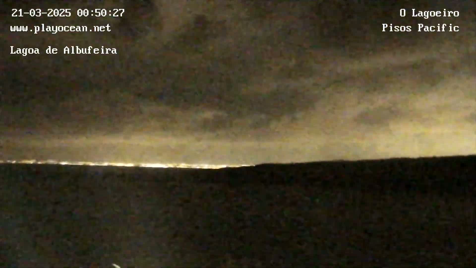 A nighttime view shows a dark landscape with a distant line of lights on the horizon under a cloudy sky.