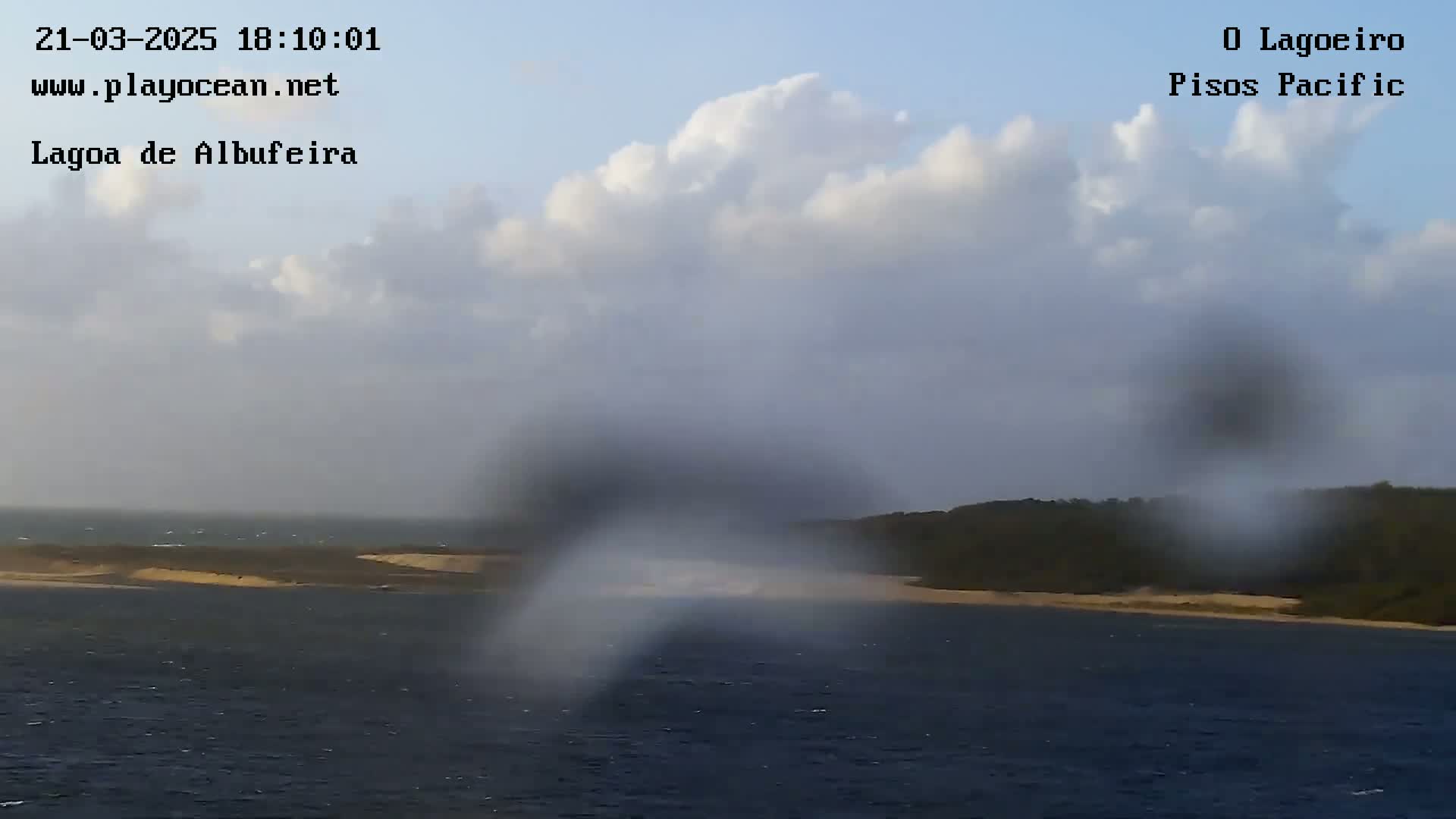 A partly cloudy day shows a body of water with a sandy shoreline and dark green vegetation beyond, viewed through a rain-streaked surface.