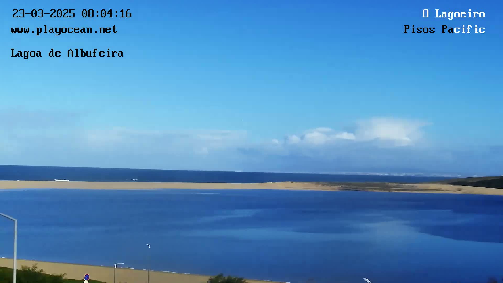 A calm, blue lagoon stretches out to a sandy shore and ocean under a mostly clear sky with a few fluffy clouds.