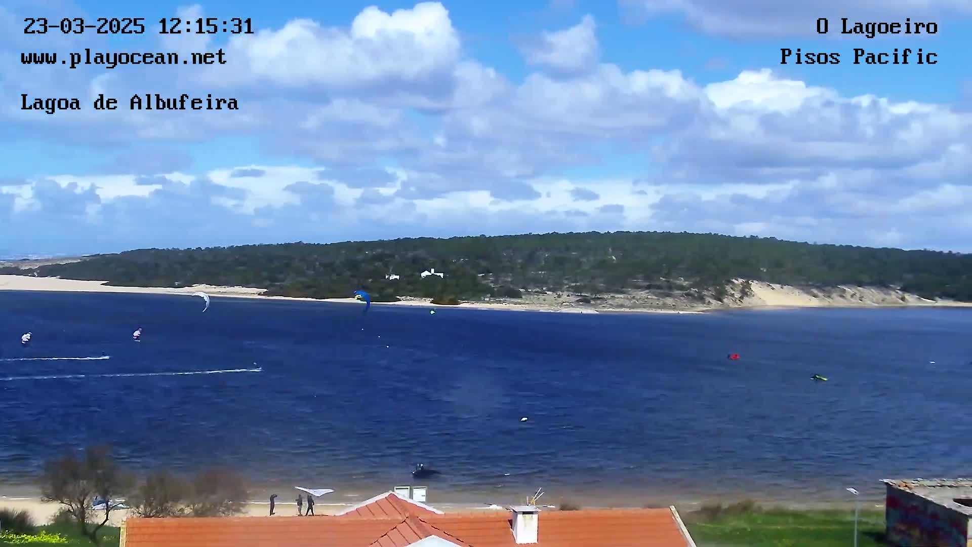 A body of water with several kitesurfers, under a partly cloudy sky, is seen from a high vantage point overlooking a sandy shore and a forested hill.