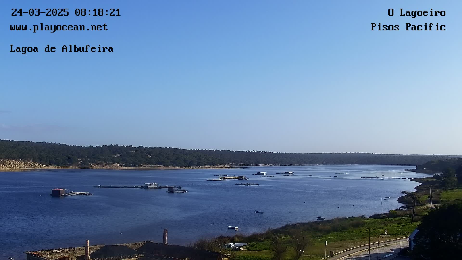 A calm body of water with several small structures and boats, surrounded by a green shoreline and a clear blue sky.