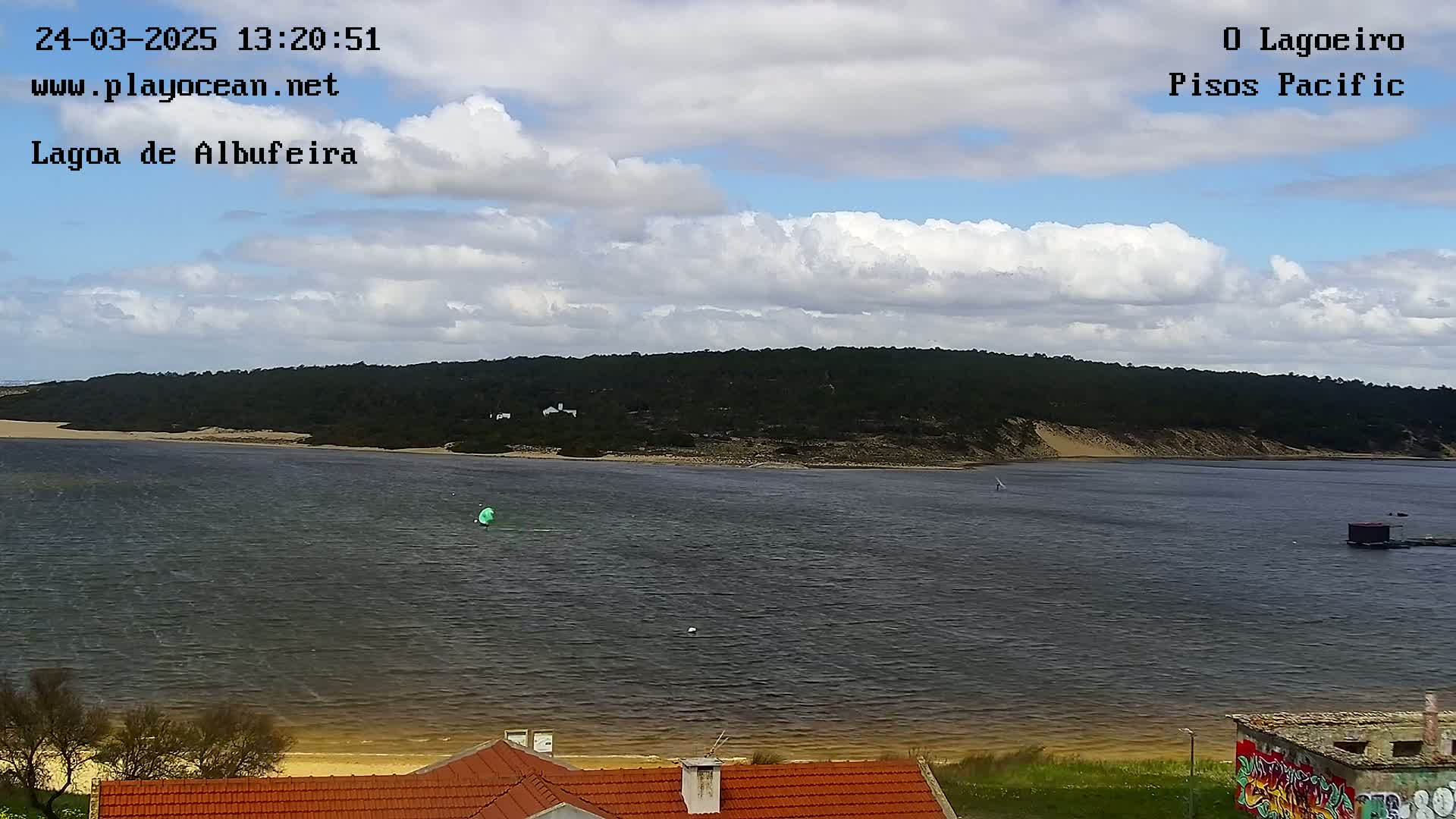 A body of water, partially sandy shore, and a verdant hillside under a partly cloudy sky are visible.