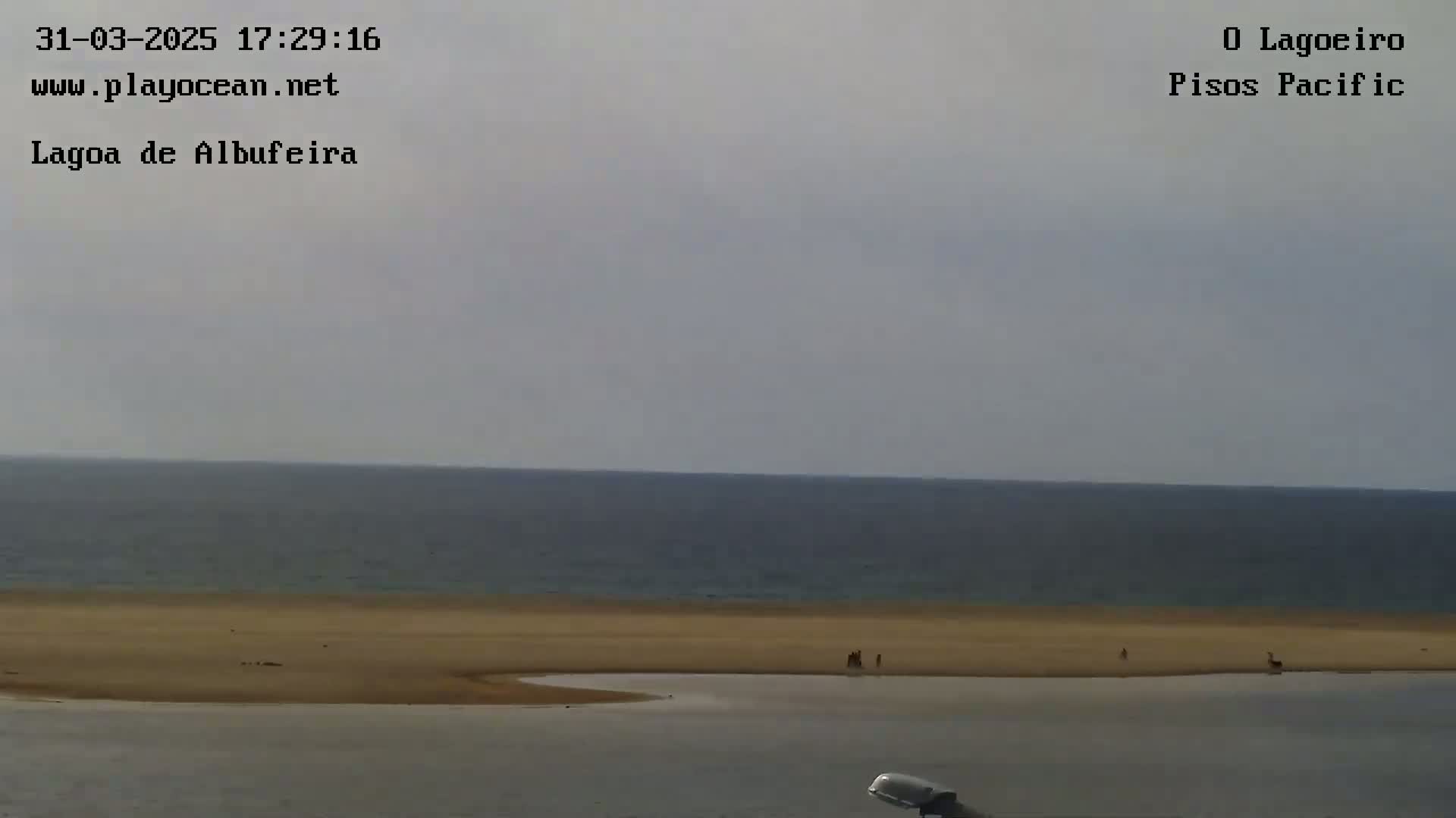 A calm ocean meets a wide, sandy beach where a few people are visible under an overcast sky.