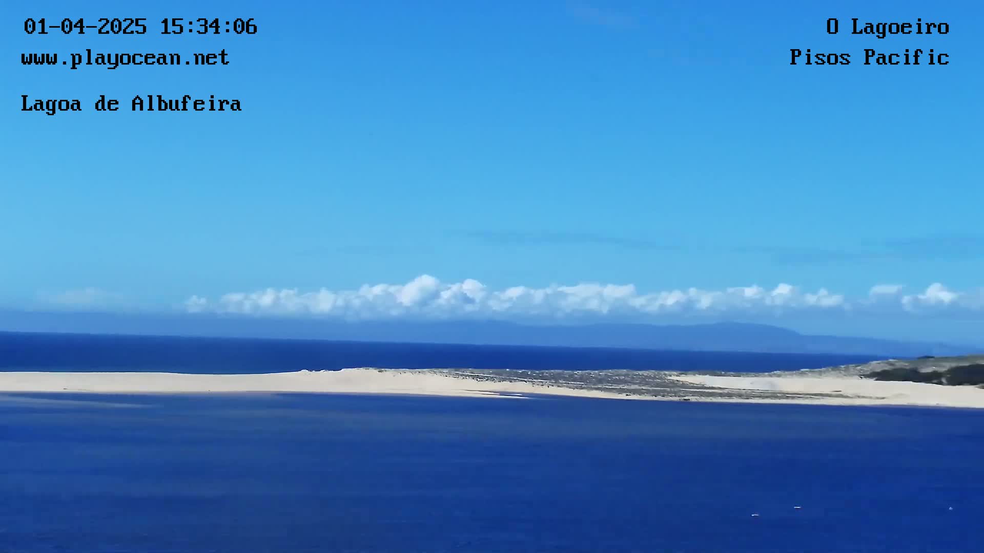 A sandy beach separates a calm, dark-blue ocean from a distant shoreline under a clear blue sky with a thin band of fluffy white clouds.