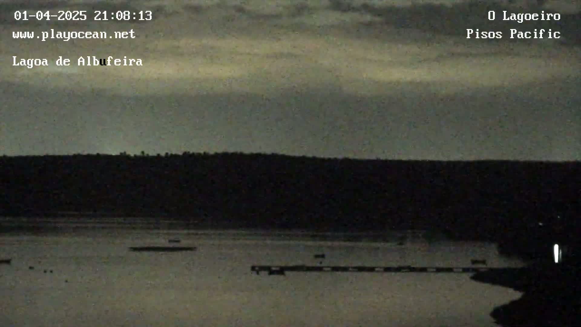 A dark, overcast sky hangs over a calm body of water with a dark shoreline and a few small objects on the water's surface.