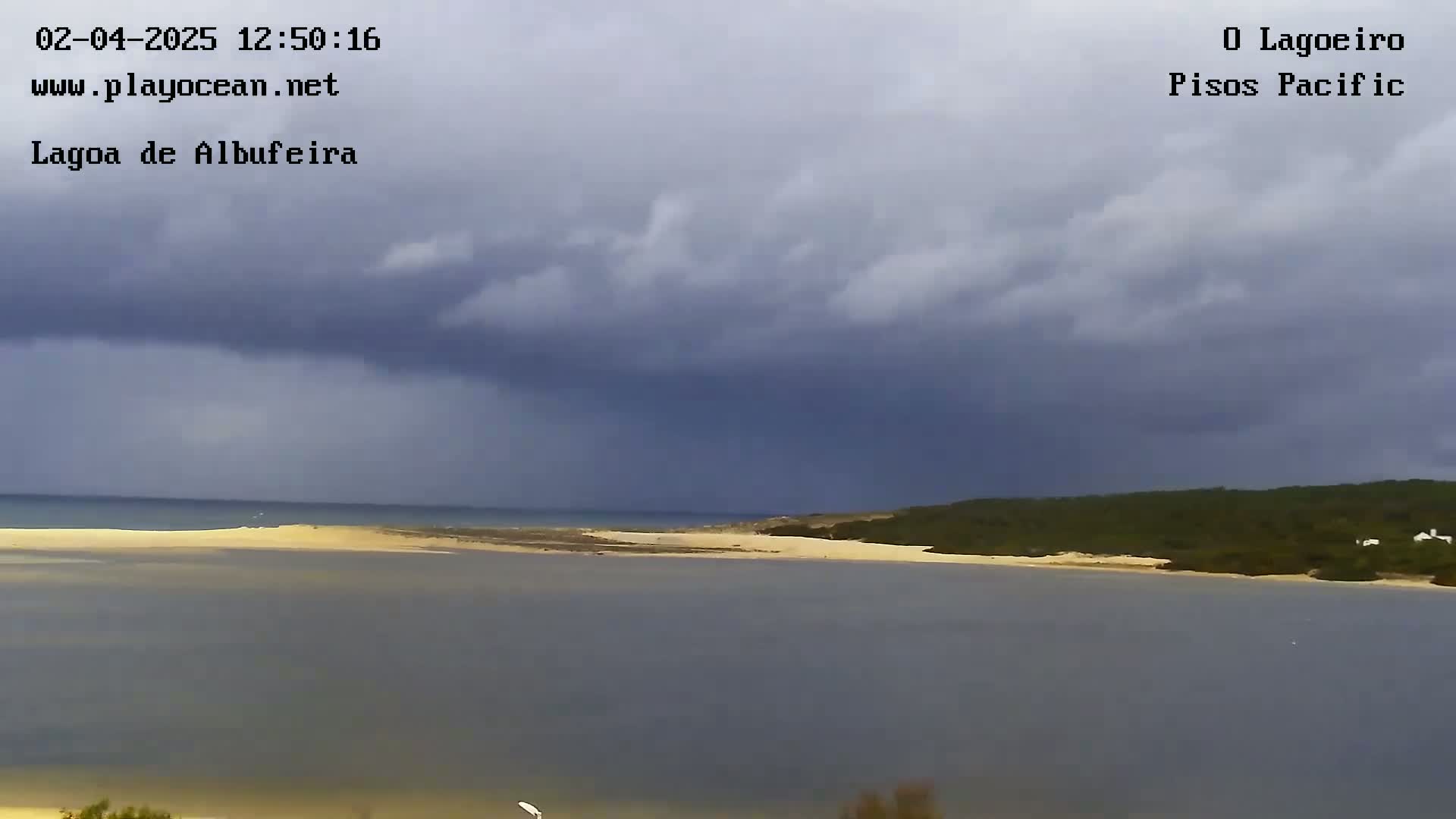 A sandy beach and calm lagoon are seen under a dark, cloudy sky.