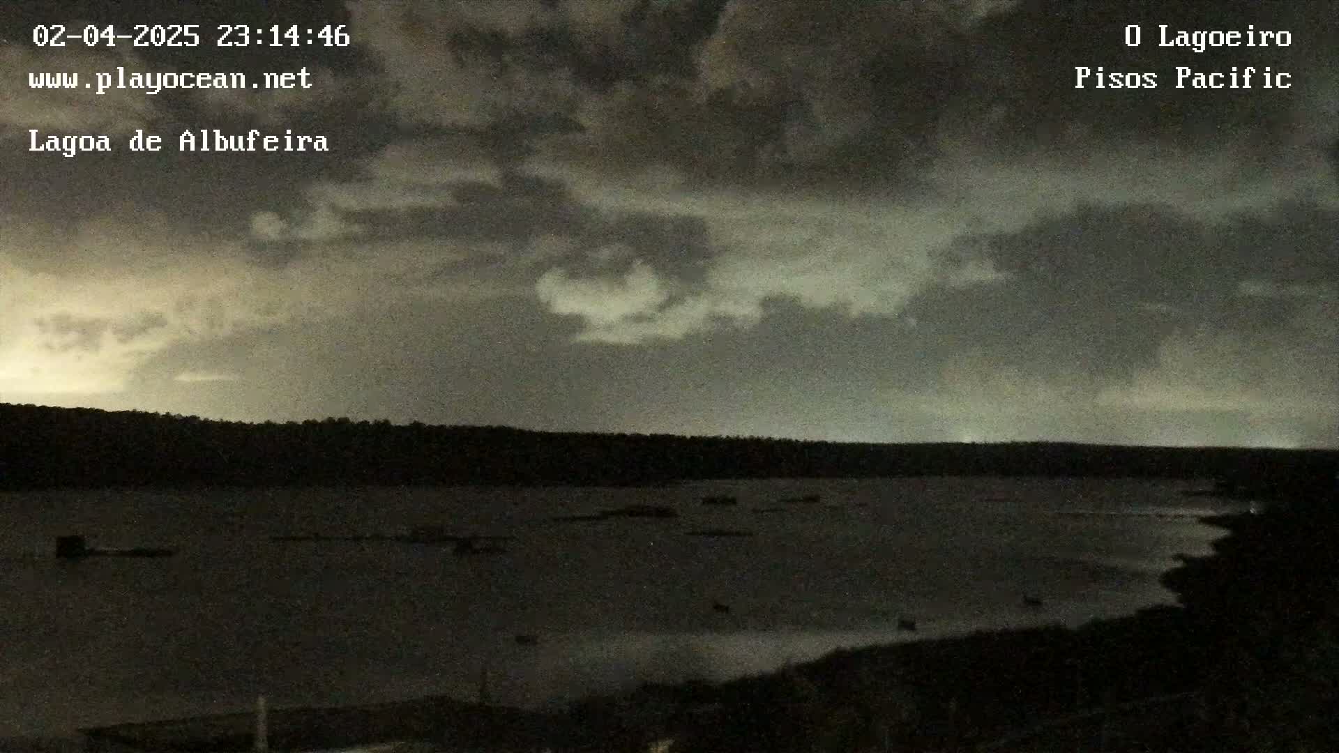 A dark, overcast night scene shows a body of water with a dark shoreline under a mostly cloudy sky.