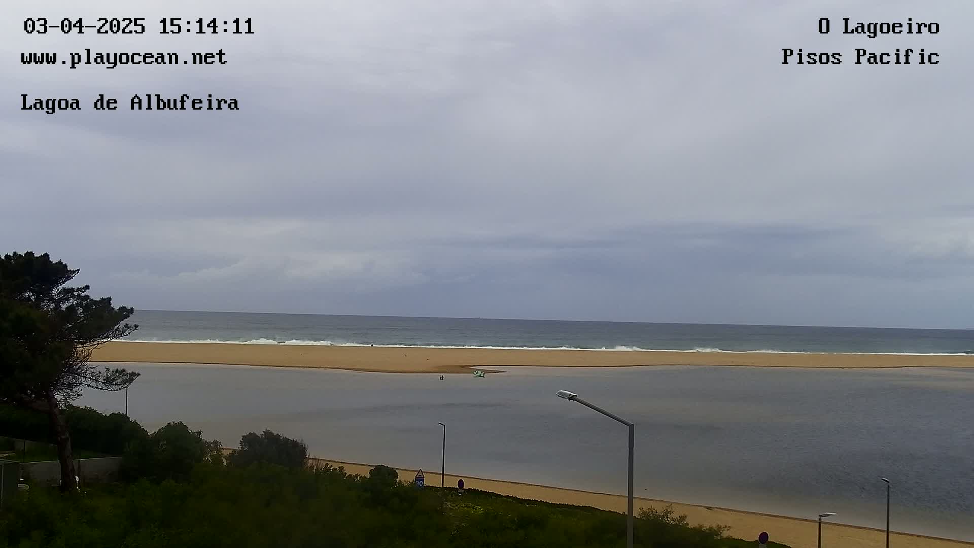 A calm lagoon meets a wavy ocean under an overcast sky, with sandy beaches and vegetation visible from a high vantage point.