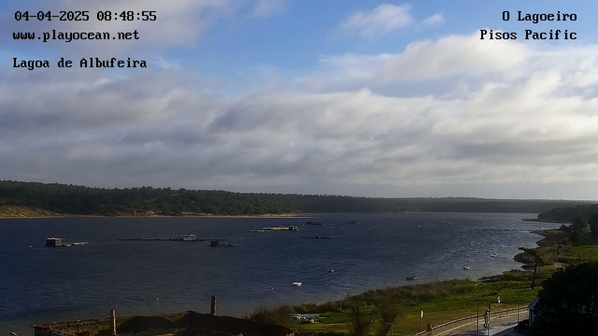A mostly cloudy day overlooks a calm body of water with several small structures and boats visible, surrounded by green vegetation and a shoreline.