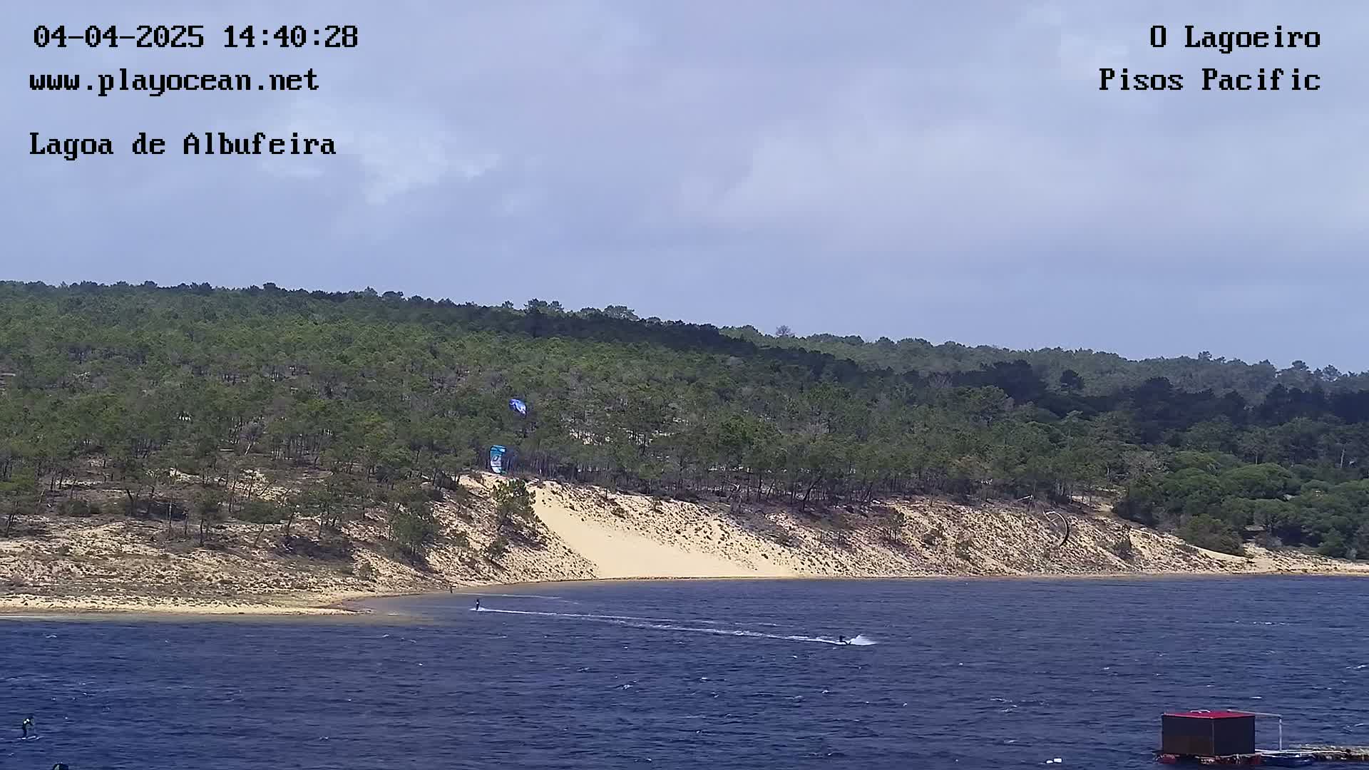 Under a partly cloudy sky, two people kitesurf on a body of water next to a sandy shoreline with a hill covered in green trees.