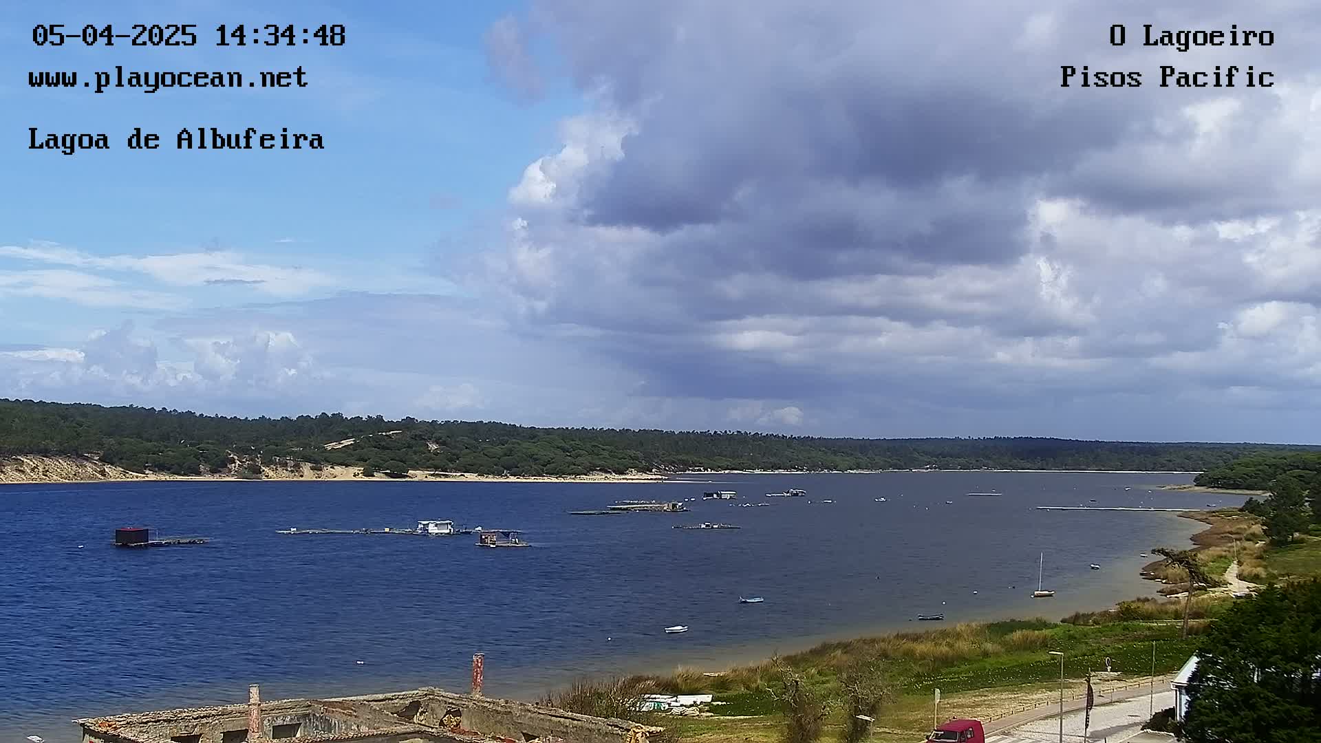 A partly cloudy sky overlooks a calm body of water with several small structures and boats visible, surrounded by green vegetation.