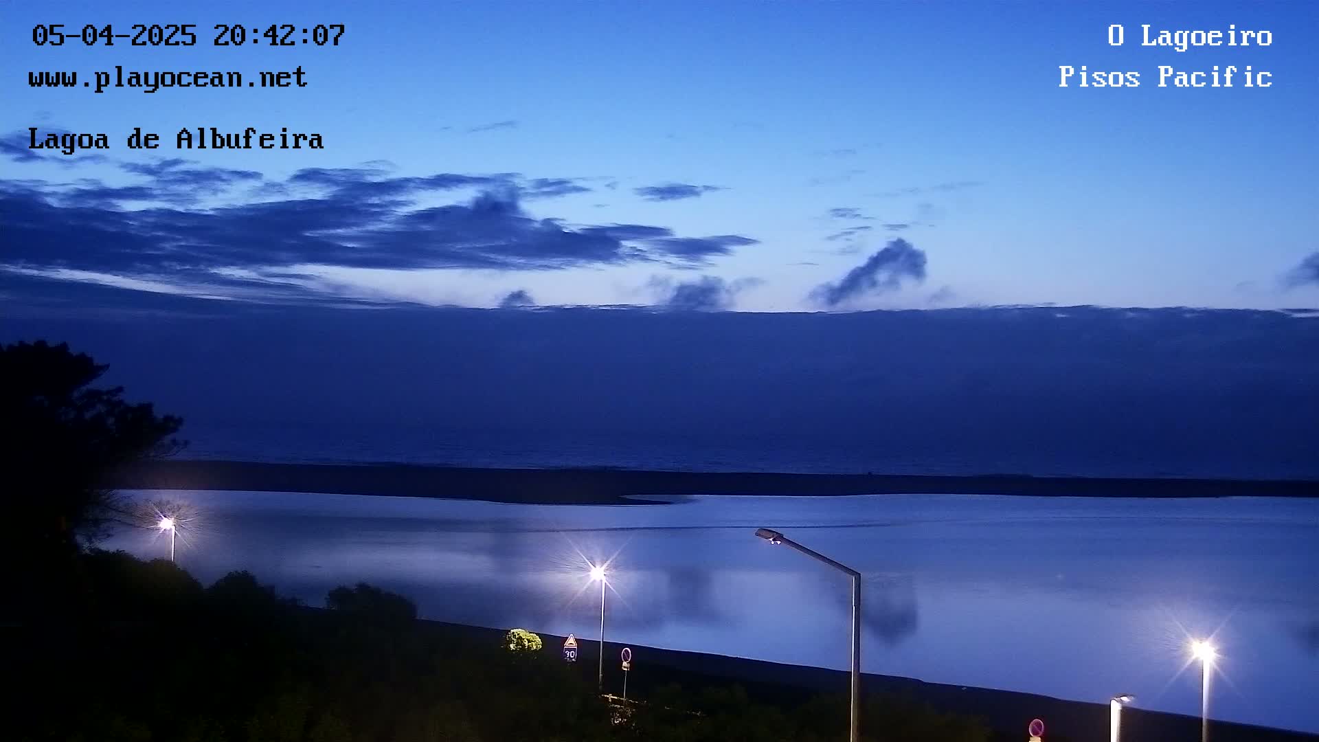 Under a twilight sky with scattered clouds, a calm body of water is illuminated by streetlights along a dark shoreline.